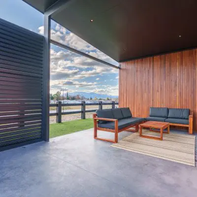 Modern patio with NSW Spotted Gum wooden walls, featuring black sofas and a wooden coffee table on a beige rug. Enjoy the view of the sky and landscape through a slatted screen crafted from exquisite Spotted Gum timber.