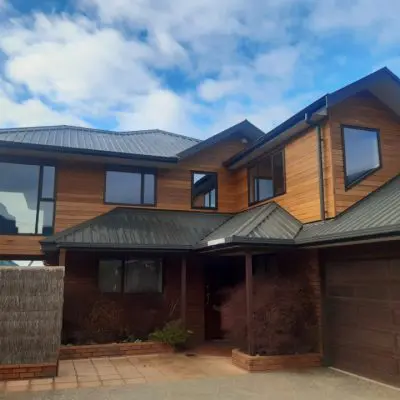 Two-story modern house with wooden siding, large windows, and a gray metal roof. Features a garage door on the right and a small front garden. Blue sky with scattered clouds in the background.