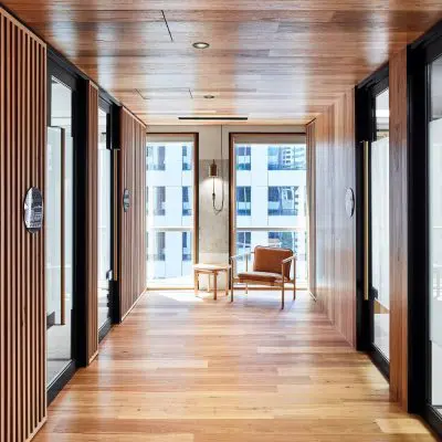 The modern hallway features shiplap timber walls complementing the wooden floors and elegant glass doors on the sides. A brown chair and small round table rest near the window at the end, with a breathtaking cityscape visible outside.