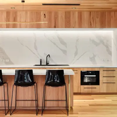 Modern kitchen featuring light wood cabinets and engineered flooring, a white marble backsplash, black chairs at an island, built-in oven, and a sleek black faucet. The elegant design is complemented by the warmth of a shiplap timber wall.