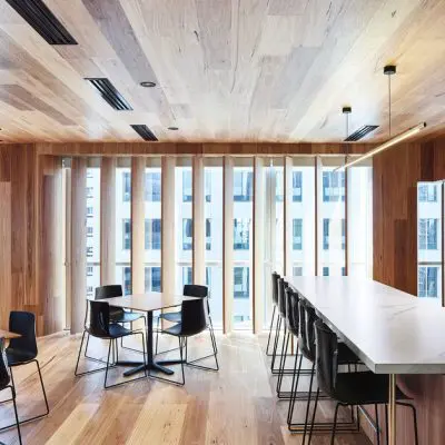 The modern office breakroom boasts shiplap timber walls and a ceiling, with sleek tables, black chairs, and a long white countertop. Large windows with vertical blinds flood the space with natural light.