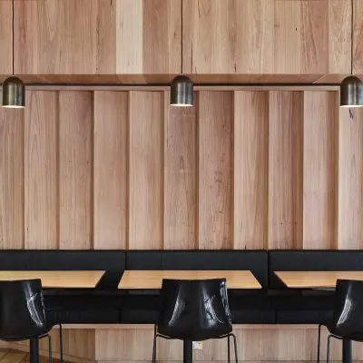 A modern cafe interior showcases a shiplap timber wall and engineered flooring, complemented by black chairs and tables. Three pendant lights hang gracefully from the ceiling.