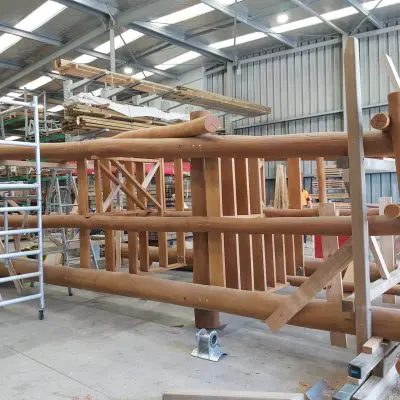 A large wooden structure, crafted from hardwood, is under construction inside a workshop. Logs and planks are supported by beams, hinting at a potential playground design. A ladder stands nearby, surrounded by various building materials in the background.