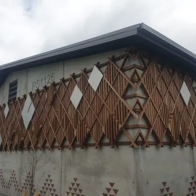 Building with a geometric wooden lattice design on its exterior and small triangular patterns on the wall. Crafted from hardwood, the structure is labeled PS 1126. Under an overcast sky, thoughtful landscaping complements its modern aesthetic.