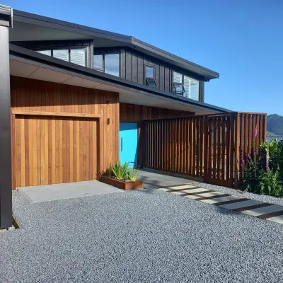 Modern house with a wooden facade and vibrant blue door, surrounded by a gravel driveway and greenery, under a clear blue sky.