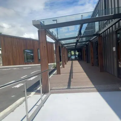 Modern building with a wooden facade and glass canopy, enhanced by sleek structural beams. A wheelchair ramp leads to the entrance, and an adjacent parking area is visible under a partly cloudy sky.