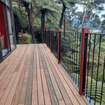 Wooden deck with black metal railing overlooks lush greenery and tree ferns. The deck is adjacent to a building with sliding glass doors.