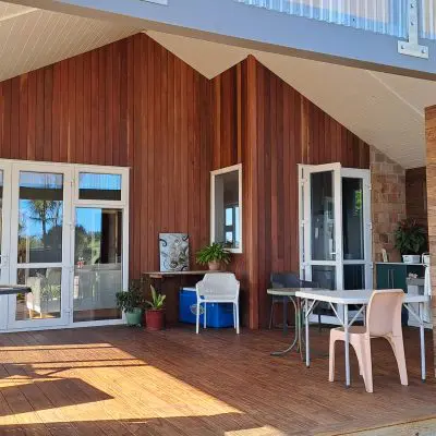 Wooden patio with a barbecue grill, table, and chairs in rich browns. Large windows and doors open to the interior. Potted plants with deep purples sit near the entrance.