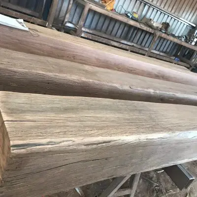 In a bustling workshop, several large wooden structural beams are carefully stacked, accompanied by posts and battens, with tools and equipment scattered in the background.