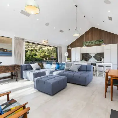 Modern open-concept living room with vaulted ceiling and engineered flooring, featuring a blue sectional sofa, dining table, and kitchen. Large windows and artwork enhance the light, airy space. A shiplap timber wall adds a touch of rustic elegance to the interior design.