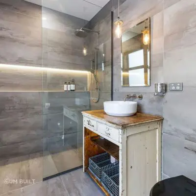 Luxurious modern bathroom with a glass-enclosed shower, wall-mounted showerhead, and an illuminated shelf. Featuring a rustic wooden vanity with a vessel sink and baskets underneath, it offers gray tiled walls and pendant lighting reminiscent of a serene escape to Okains Bay.