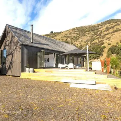 A luxury modern house with a sloped roof and large windows sits on a wooden deck, nestled in the serene landscape of Okains Bay. Surrounded by dry grass and rolling hills under a blue sky, this stunning bach offers an idyllic escape.