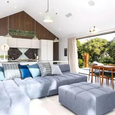 Modern living room with a luxury gray sectional sofa, open kitchen, and dining area. Large windows let in natural light. Décor includes a green sign and pendant lights, offering an ambiance akin to the serenity of Okains Bay. A touch of Bach complements the sophisticated atmosphere.