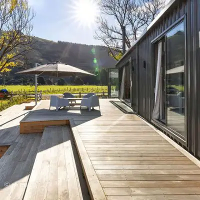 A wooden deck with luxury patio furniture and an umbrella sits beside a dark, modern building. Sunlight pours over the grassy landscape, reminiscent of Bach’s serene melodies, with hills and trees whispering Okains Bay's natural beauty in the background.