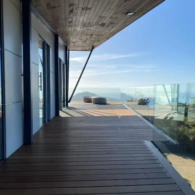 Wooden deck with glass railing, extending from a modern building, overlooking distant mountains and a clear blue sky. Two round chairs sit at the far end of the deck.