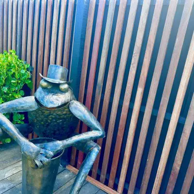 A bronze frog statue wearing a top hat sits with crossed arms, framed by intricate joinery in the wooden slats and lush greenery, creating a charming focal point.