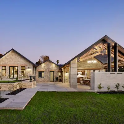 Modern house with stone exterior and large patio, featuring a covered outdoor space with exposed beams and elegant trusses. It is set against a backdrop of trees and a sunset sky.