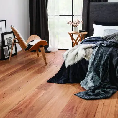 A modern bedroom with engineered wooden flooring features a bed with rumpled dark bedding, a solid wooden chair, framed art on the floor, and a side table with flowers by a large window.