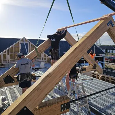 Construction workers install large hardwood beams and trusses on a building site, using ladders and safety equipment under a clear sky.