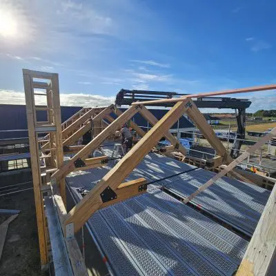 Hardwood trusses are under construction at a building site, framed by scaffolding and a crane in the background on a sunny day.