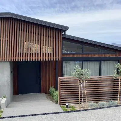 A modern house with wooden slats made of Queensland Spotted Gum, large windows, and a blue door. It features a small fence and young trees at the entrance.