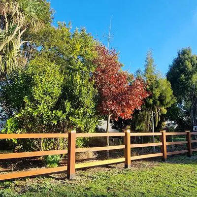 A hardwood fence with vertical posts and horizontal rails elegantly borders a grassy area adorned with various trees, including one with vibrant red leaves, all under a clear blue sky, creating a picturesque landscaping scene.