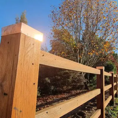 A hardwood fence with metal-topped posts in the foreground extends into the distance, showcasing fine landscaping. Sunlight reflects off the metal, enhancing the warm hues of autumn foliage against a clear blue sky in the background.