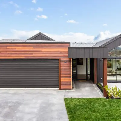 Modern house with a blend of wooden, brown accents and dark materials, featuring a large garage door, glass front entry, and surrounding lawn.