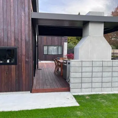 The modern house exterior features dark wood siding treated with clear oil, complemented by concrete and block walls. An outdoor dining area with wooden chairs overlooks a lush green lawn in the foreground, creating an inviting and stylish atmosphere.