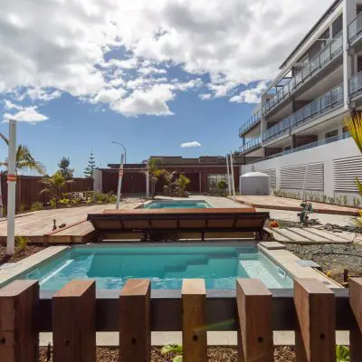 View of two small pools in a residential complex with hardwood decking, surrounded by lush landscaping and a modern building.