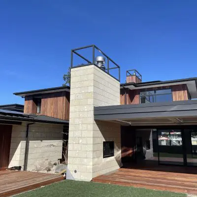 Modern two-story house with hardwood paneling, large glass windows, and a central stone chimney. The house features a manicured lawn adorned with an Ironbark tree and a spacious wooden deck under a clear blue sky.