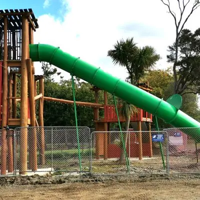 The tall playground structure, featuring a green tube slide and encircled by a chain-link fence, stands proudly beside towering hardwood trees under a cloudy sky.