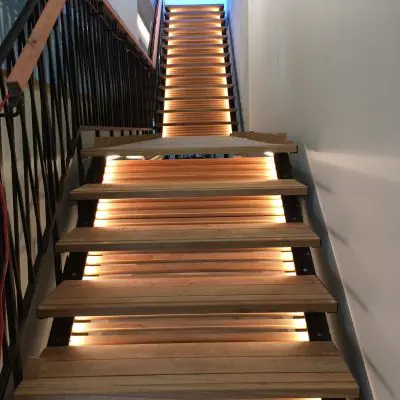 A stunning wooden staircase with solid, engineered steps and built-in LED lights beneath each one. This architectural marvel is flanked by a sleek black metal railing as it gracefully ascends to the upper floor.