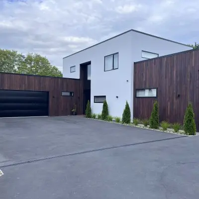 Modern house with a white and dark wood exterior, featuring large sawn windows and a spacious driveway adorned with greenery. A cloudy sky forms the perfect backdrop, enhancing the home's clear oil finish's elegance.