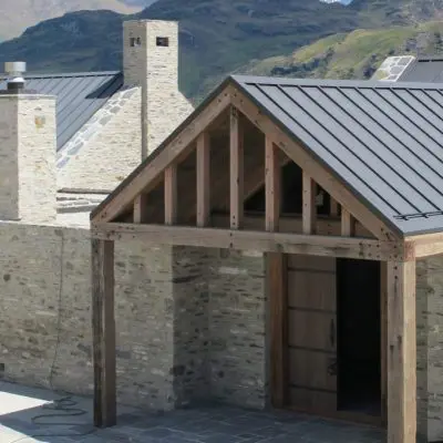 Stone building with wooden and metal roof, featuring a gabled entrance supported by sturdy posts. Mountainous landscape is visible in the background.