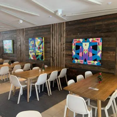 A dining area with wooden tables and white chairs. Colorful abstract paintings are on the wooden walls. Small plants decorate the tables.