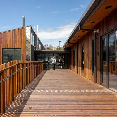 Wooden walkway with railing leading to a building entrance. Two people are walking toward the doorway under a clear blue sky.