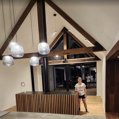 A person stands in a modern room, where trusses enhance the high vaulted ceiling with exposed wooden beams and a row of pendant lights illuminates the hardwood counter below.