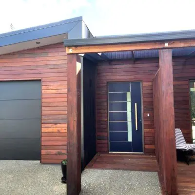 Modern house entrance with dark wood paneling and sleek battens, a large blue door with glass accents, and a connected garage. The patio features a chair strategically placed near the entrance, creating an inviting atmosphere.