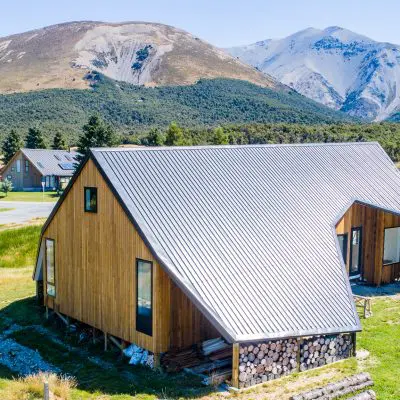 A modern wooden house, crafted using ACQ-treated materials, boasts a sloped metal roof and is enveloped by mountains and greenery. In the background, other houses and a winding road are visible.