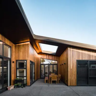 Modern house exterior with wooden paneling and large glass doors. A central patio features a wooden table and chairs, and a skylight is visible in the sloped roof.