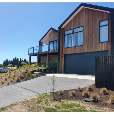 Two modern wooden houses with large windows and a shared driveway are situated on a landscaped slope under a clear blue sky.