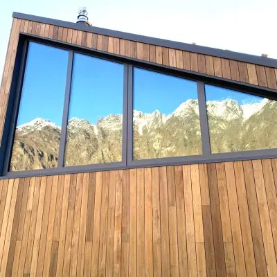 Wooden building with large angled windows reflecting snow-capped mountains under a clear sky.