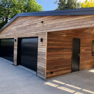 Wooden garage with two black doors, a side door, and a window, set against a backdrop of trees and a clear sky.