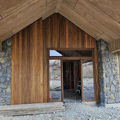 A wooden house under construction, featuring elegant stone accents and the rich hues of NSW Spotted Gum. The central entrance is open, revealing the intricate interior framework. Inside, a ladder is surrounded by construction materials under an overcast sky.