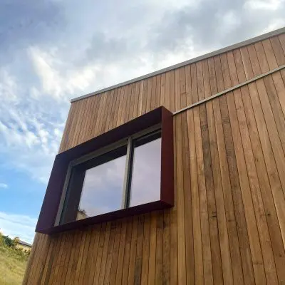 The wooden building exterior, finished with mixed pales and sawn with oil, features a large rectangular window set against a partly cloudy sky.