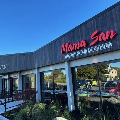 The exterior of the building, showcasing signage for "Mama San: The Art of Asian Cuisine" and "M.M Linen," features large windows framed in sawn with oil-treated wood and a mixed pales motif, all overlooking a spacious parking lot.