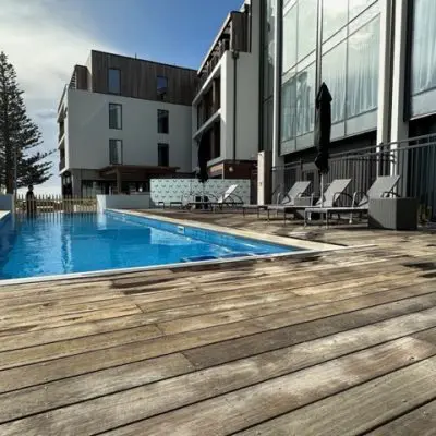 An outdoor pool with wooden decking in a lightly brushed finish is surrounded by lounge chairs and a fence. A modern multi-story building stands in the background under a partly cloudy sky.