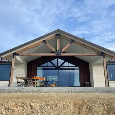 A contemporary house with structural beams, large windows, and a gabled roof. There's a patio featuring outdoor furniture and a dog sitting near the entrance.