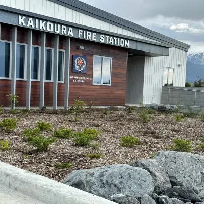 The Kaikoura Fire Station, accented with Red Ironbark wooden panels, showcases windows and a sign with a fire emblem. In the foreground, shrubs and rocks are thoughtfully arranged, all set against the majestic backdrop of towering mountains.